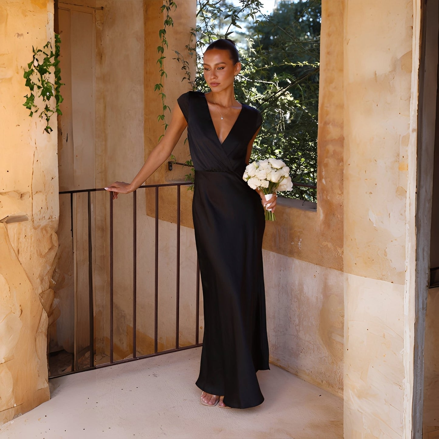 Woman in a black dress holding flowers on a balcony with stone walls and greenery.
