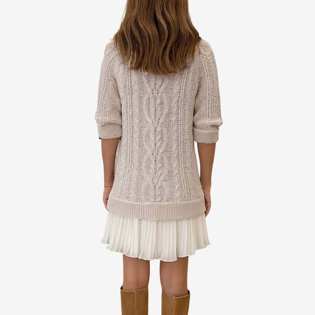 Person wearing a beige cable knit sweater over a white pleated skirt on a white background