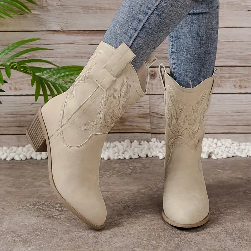Beige cowboy boots with embroidery worn by a person on a wooden floor.