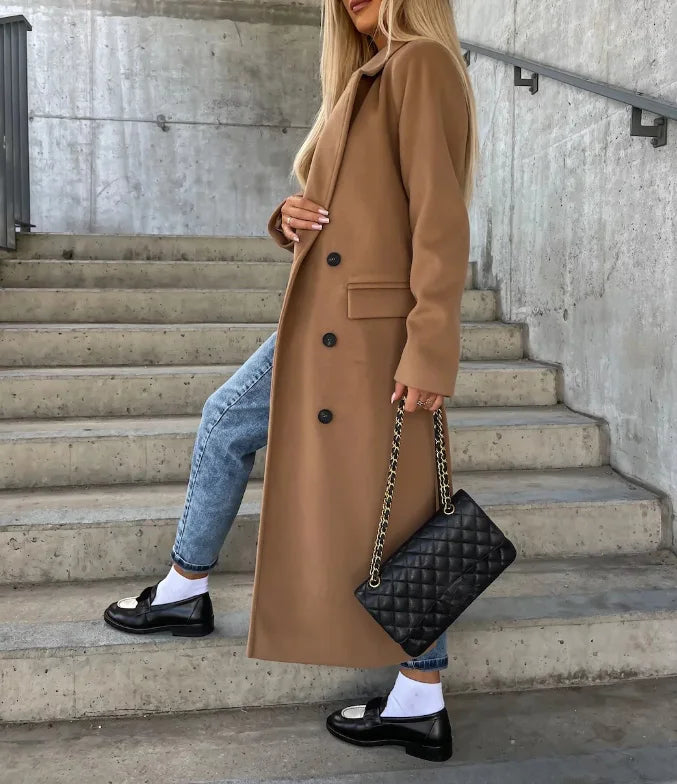 Person wearing a long brown coat and holding a black quilted handbag on concrete steps.