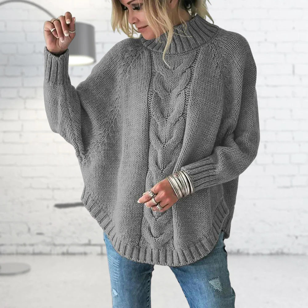 Person wearing a gray cable knit sweater with a white brick wall background