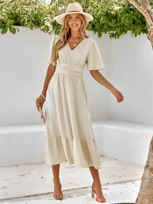 Woman in a beige dress and hat standing in a sunlit outdoor setting with greenery.