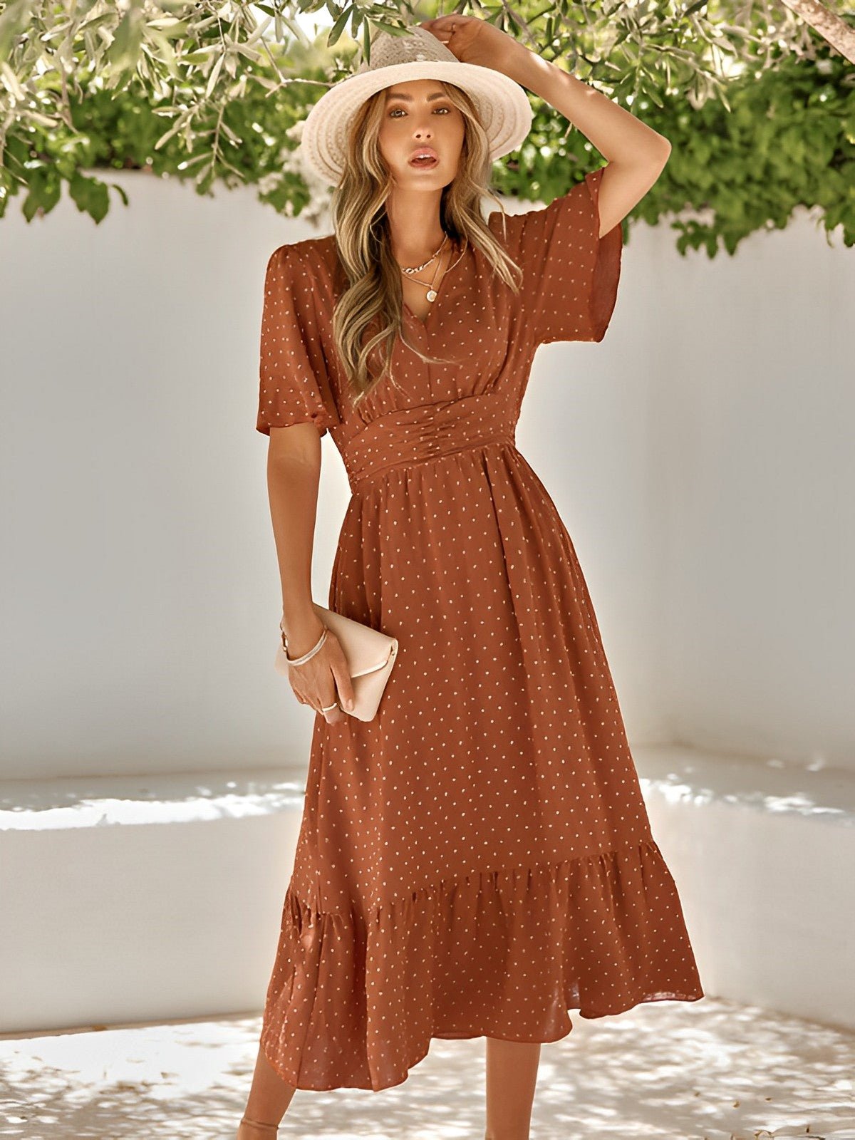 Woman wearing a rust-colored polka dot dress with a white hat, standing against a white wall with greenery.