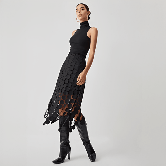 Woman wearing a black sleeveless top and black patterned skirt with black knee-high boots on a light gray background