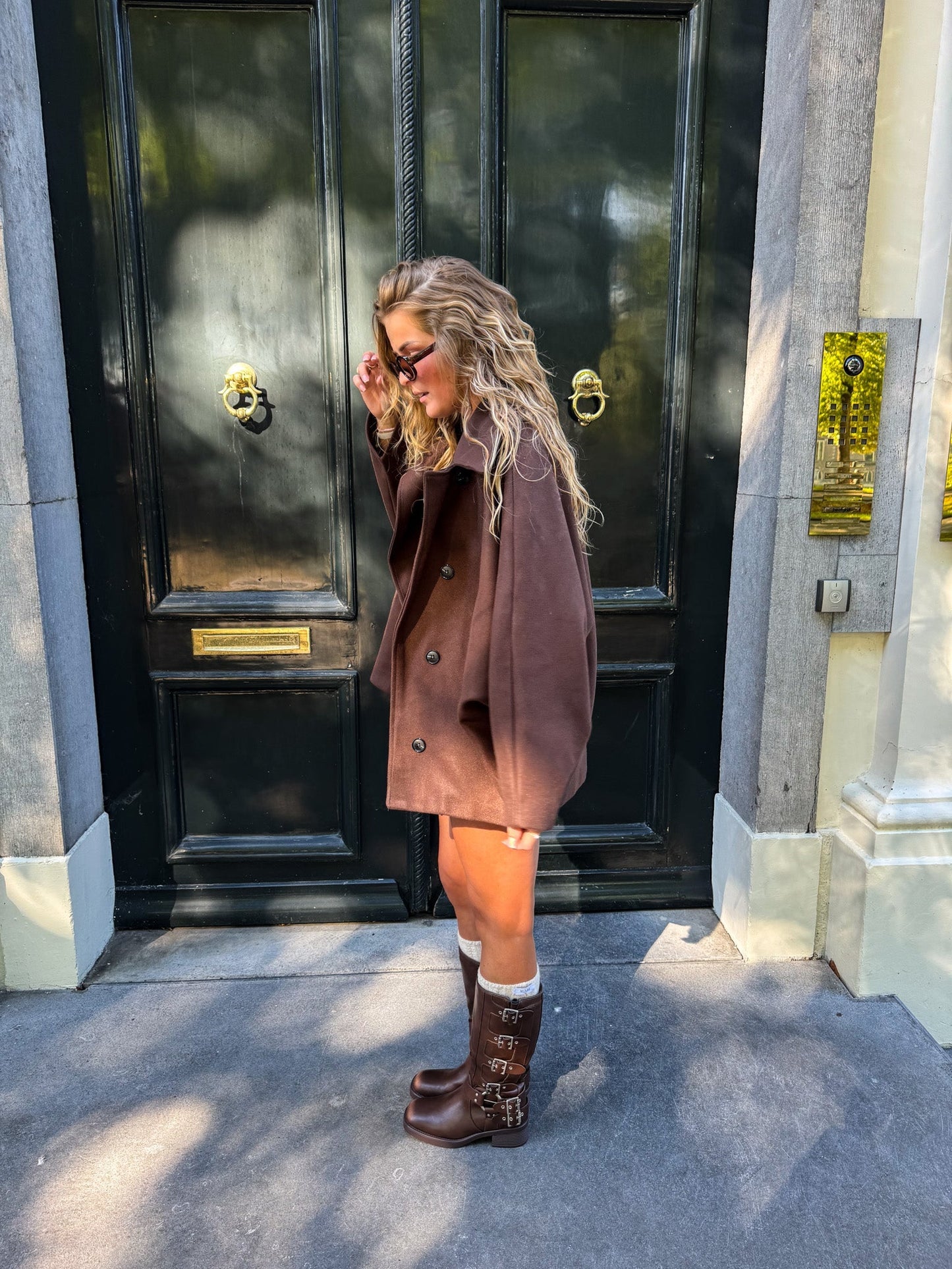 Woman in a brown coat and boots standing in front of a black door.