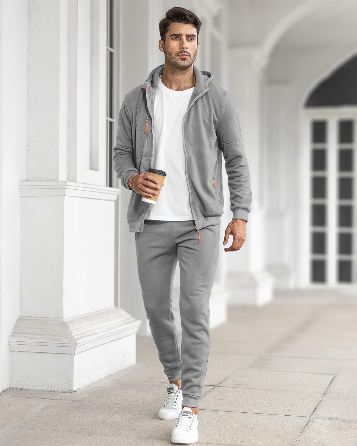 Man wearing a gray tracksuit with a white shirt, holding a coffee cup, standing in front of a white building.