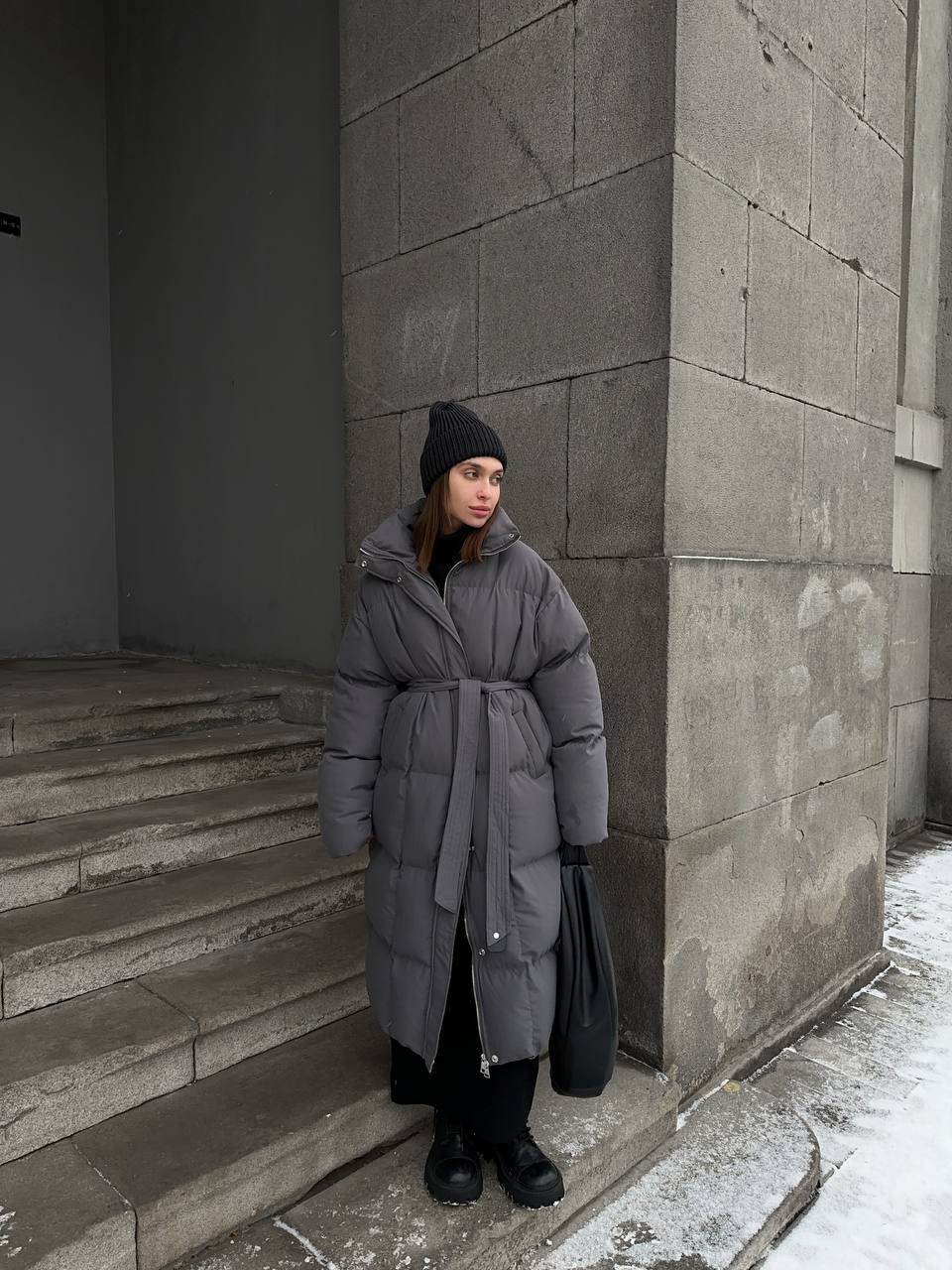 Person wearing a long gray puffer coat standing on stone steps against a gray wall.
