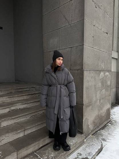 Person wearing a long gray puffer coat standing on stone steps against a gray wall.