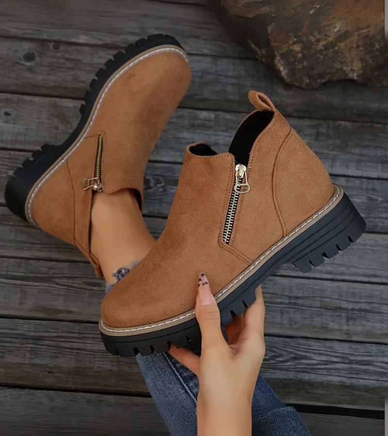 Brown ankle boots with zippers held by a hand on a wooden floor.