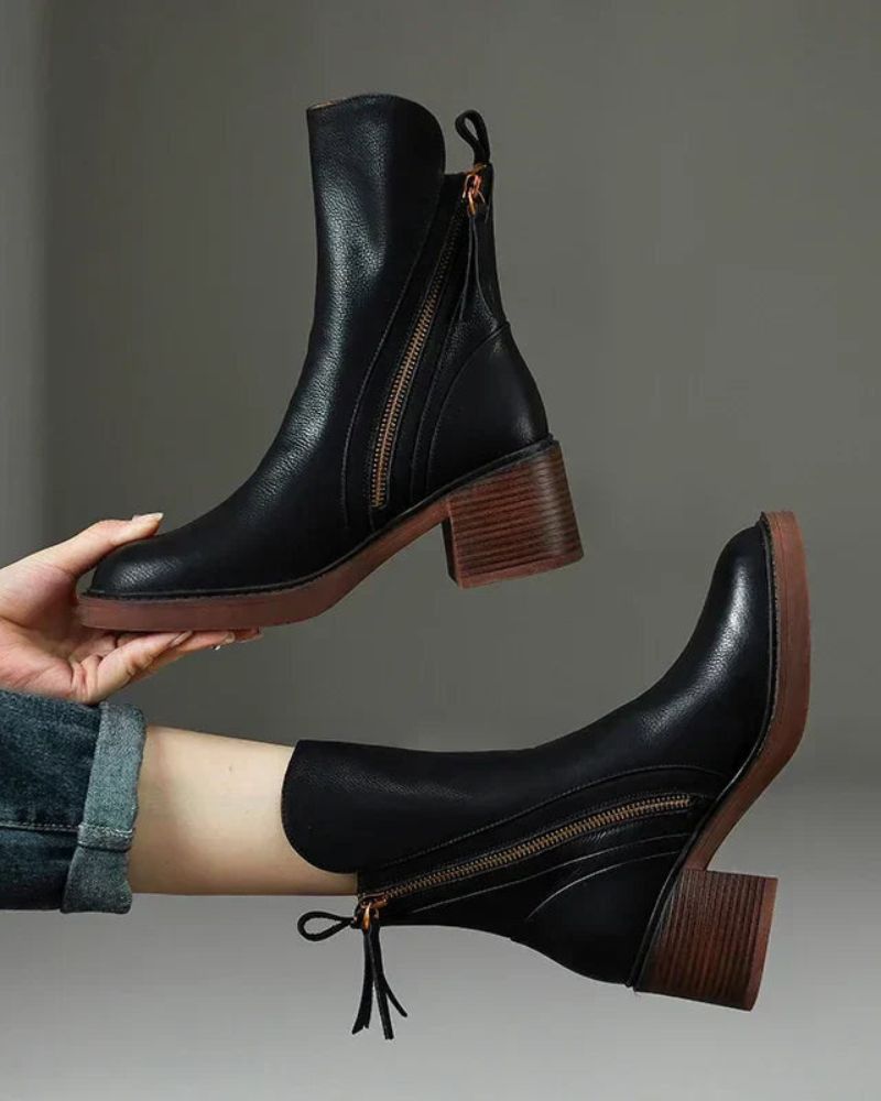 Black leather ankle boots with brown soles held by a hand against a gray background