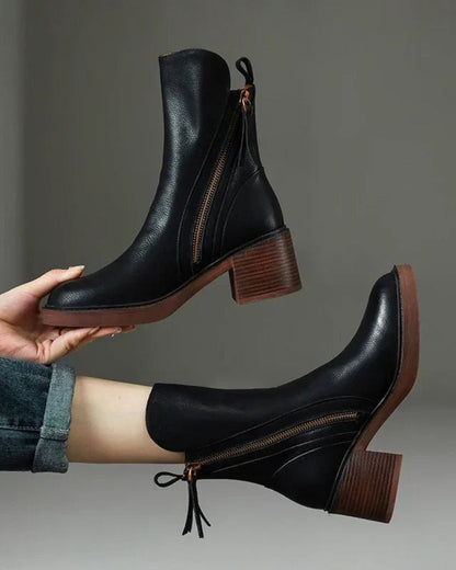 Black leather ankle boots with brown soles held by a hand against a gray background