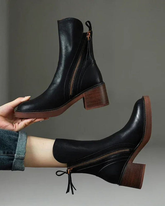 Black leather ankle boots with brown soles held by a hand against a gray background