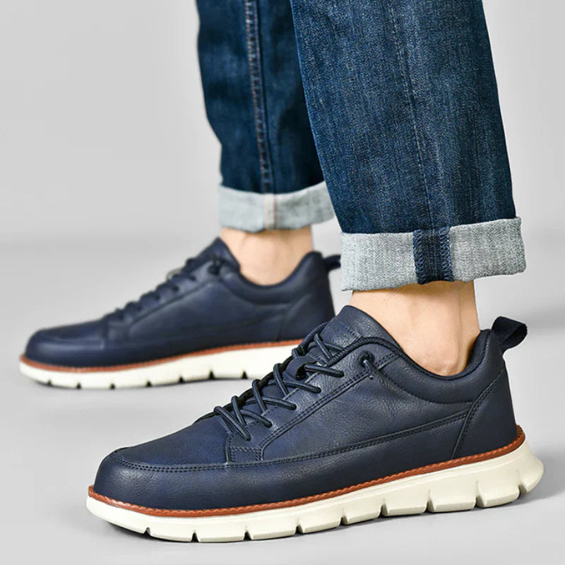 Navy blue leather shoes with white soles worn with rolled-up jeans on a gray background