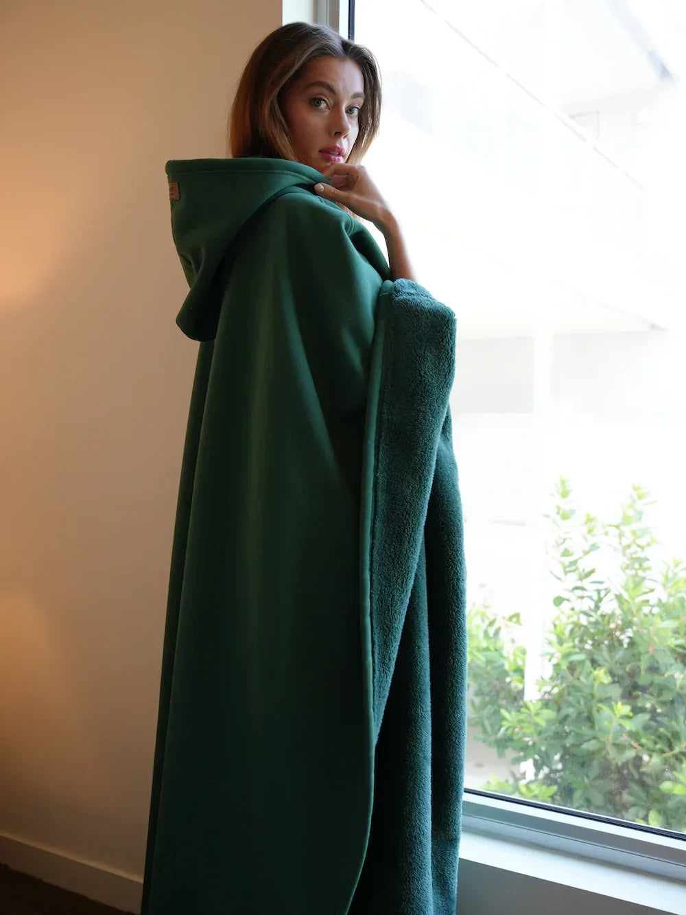 Person wearing a green hooded cloak standing by a window with a view of trees.
