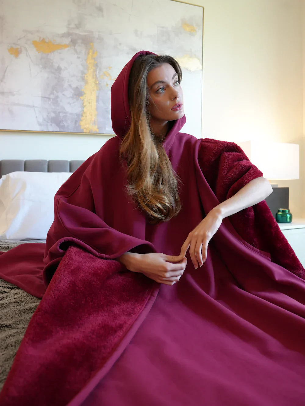 Woman wearing a burgundy hooded cloak in a bedroom setting