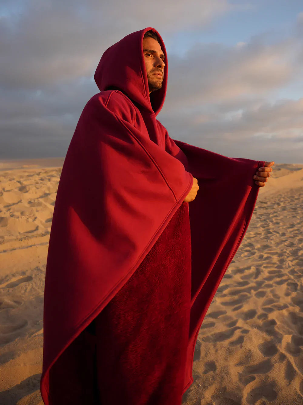 Man wearing a red hooded cloak standing in a desert landscape with a cloudy sky.