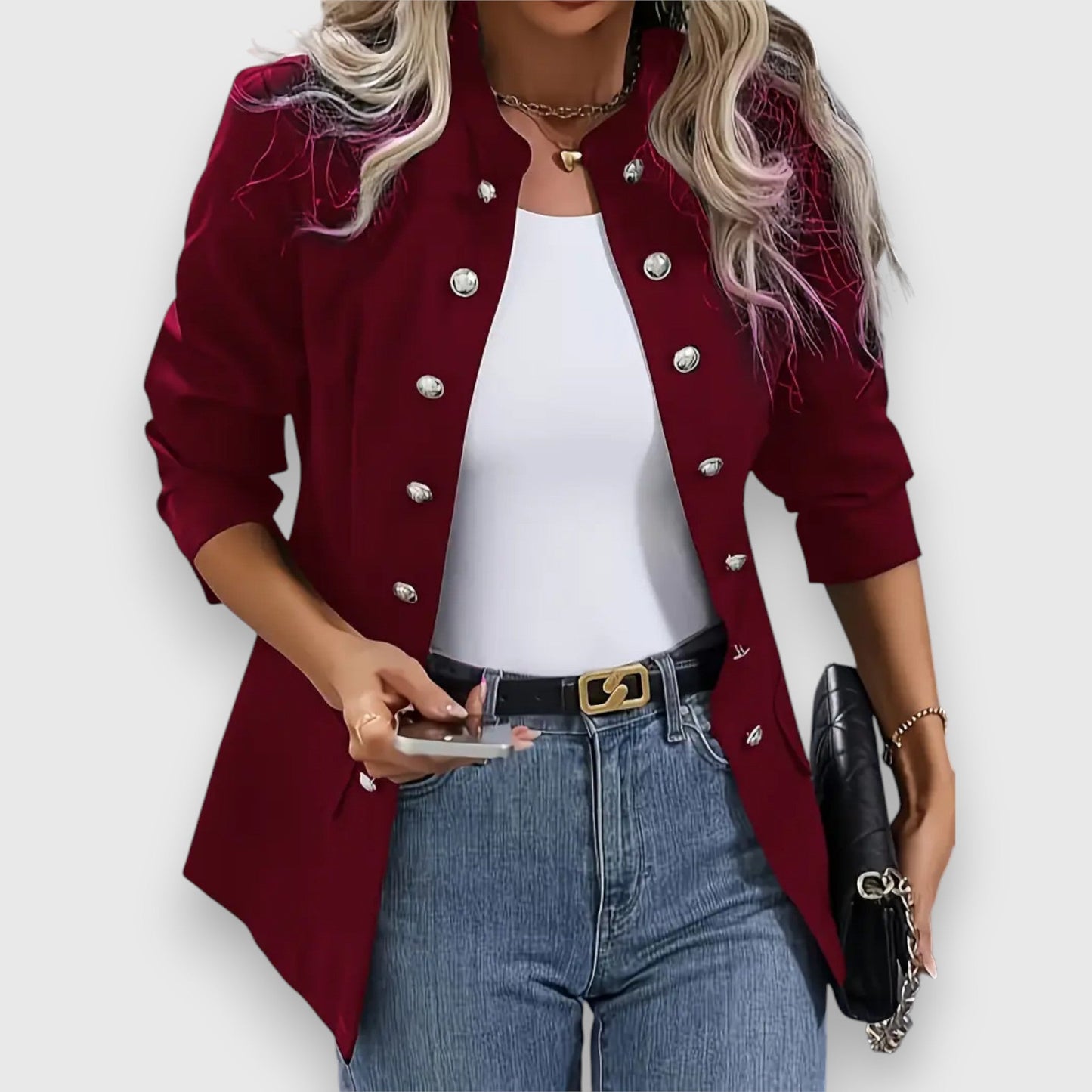 Woman wearing a red blazer with silver buttons, white top, blue jeans, and holding a phone and black clutch.