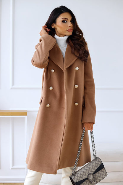 Woman wearing a brown coat with white buttons, holding a black handbag indoors.