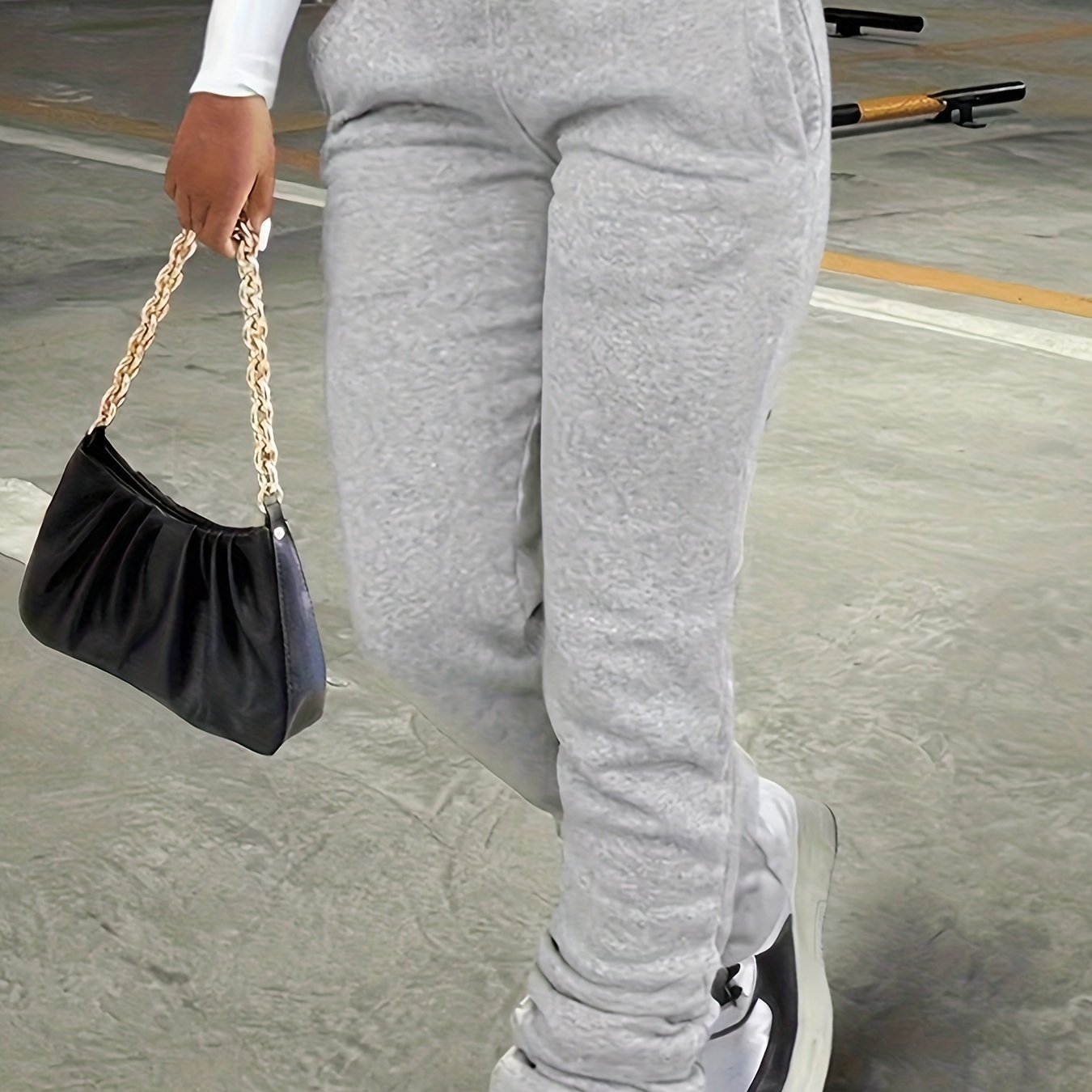 Person wearing gray sweatpants holding a black handbag with gold chain strap on a concrete floor.