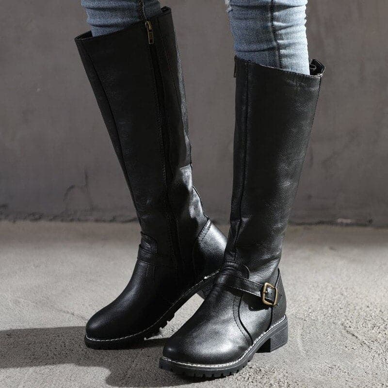 Black knee-high boots with buckles worn by a person on a gray background