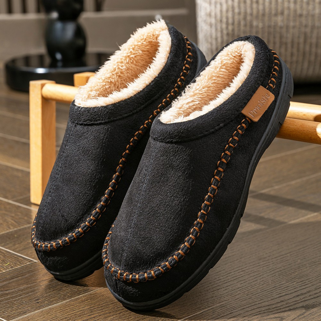 Pair of black slippers with brown stitching on a wooden floor.