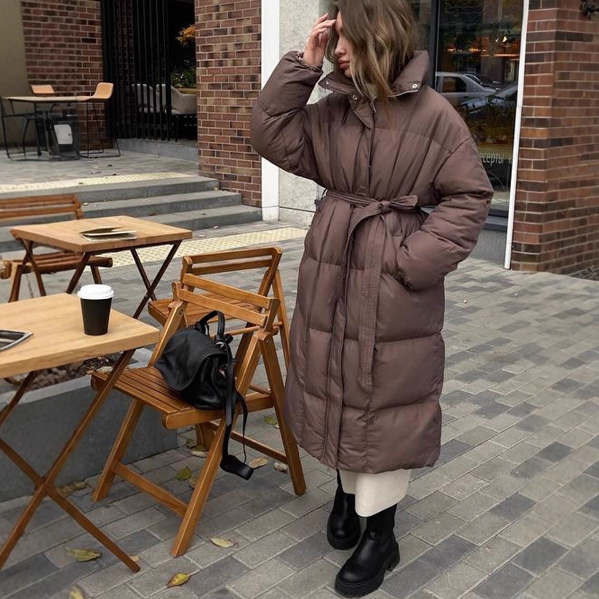 Woman in a long brown puffer coat standing outdoors near wooden chairs and tables.