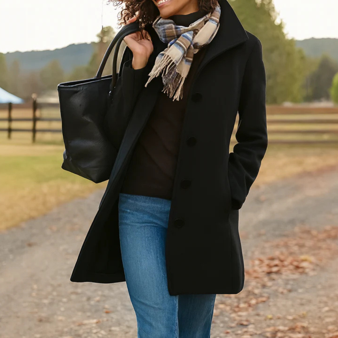 Woman wearing a black coat, blue jeans, and a plaid scarf, holding a black handbag outdoors.