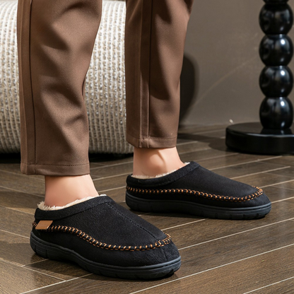 Person wearing black slippers with brown trim on a wooden floor.