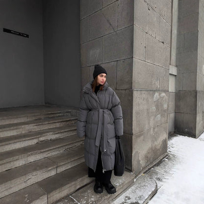 Person wearing a gray coat and black beanie standing against a concrete wall with snow on the ground.