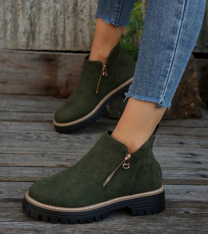 Green suede ankle boots with gold zippers worn by a person on wooden steps.