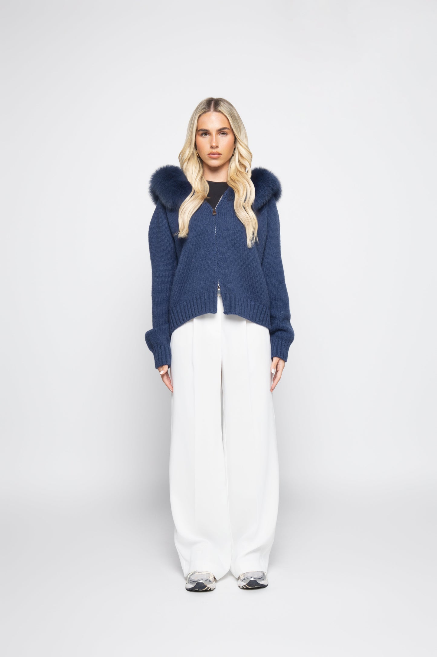 Woman wearing a blue hoodie with fur hood and white pants on a white background