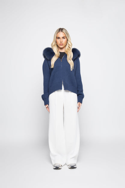 Woman wearing a blue hoodie with fur hood and white pants on a white background