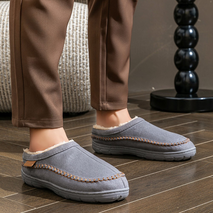 Person wearing gray slippers with brown trim on a wooden floor.