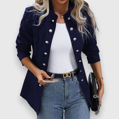 Woman wearing a navy blue blazer with silver buttons, white top, and blue jeans on a light gray background.