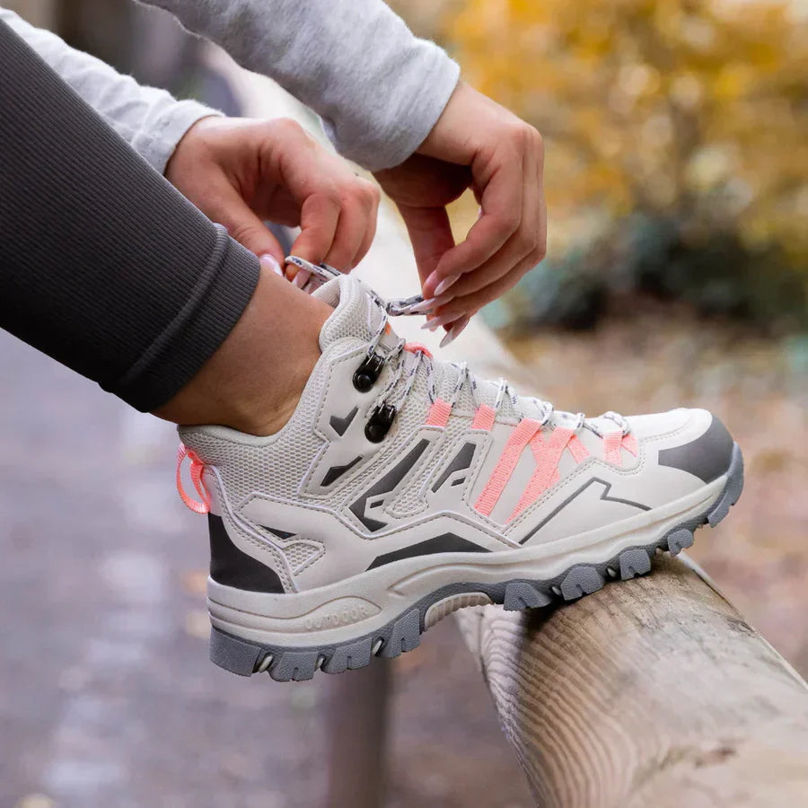 Person tying the laces of a white hiking shoe with pink accents outdoors.