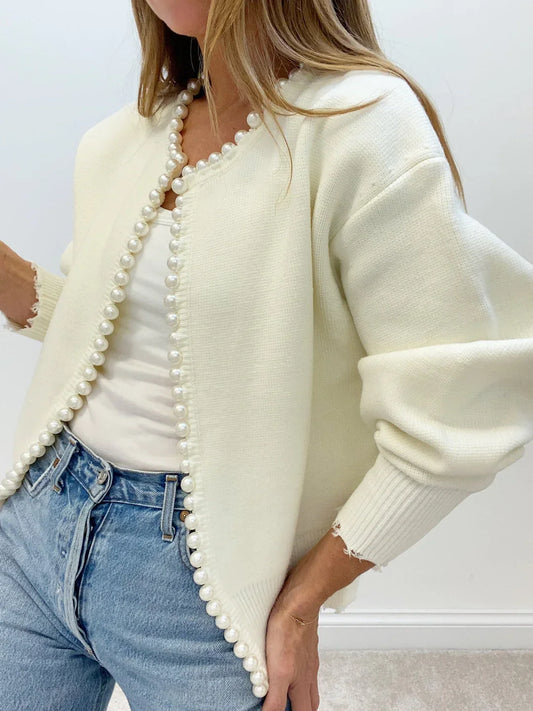 Woman wearing a cream cardigan with pearl details, white top, and blue jeans.