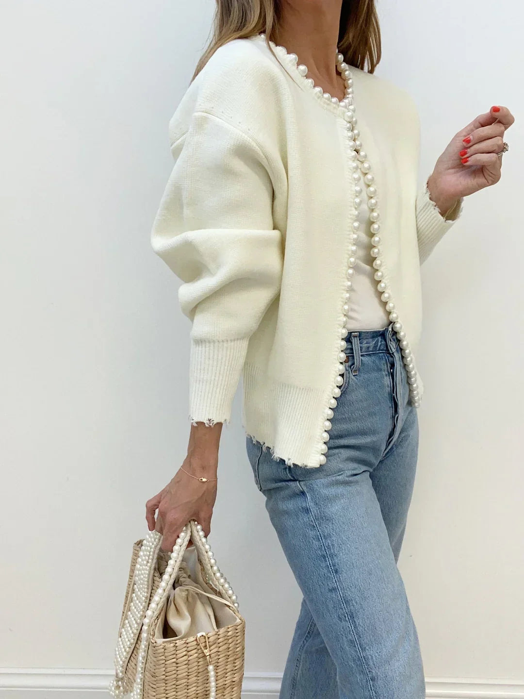 Woman wearing a cream cardigan with pearl trim and holding a straw bag against a white background
