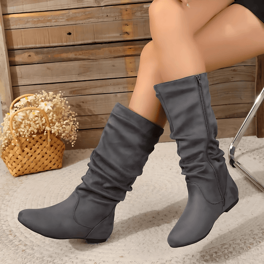 Gray knee-high boots worn by a person sitting on a carpeted floor with a wooden wall in the background.