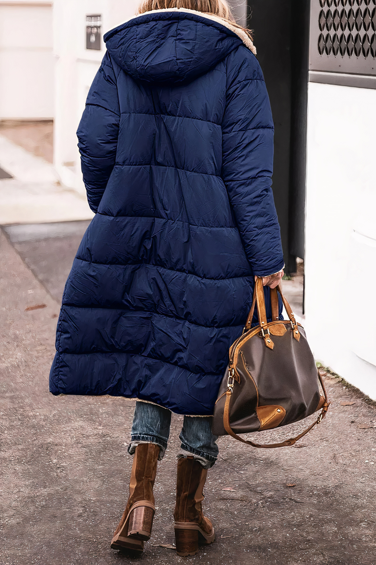 Person wearing a long blue puffer coat with a hood, holding a brown handbag on a street.