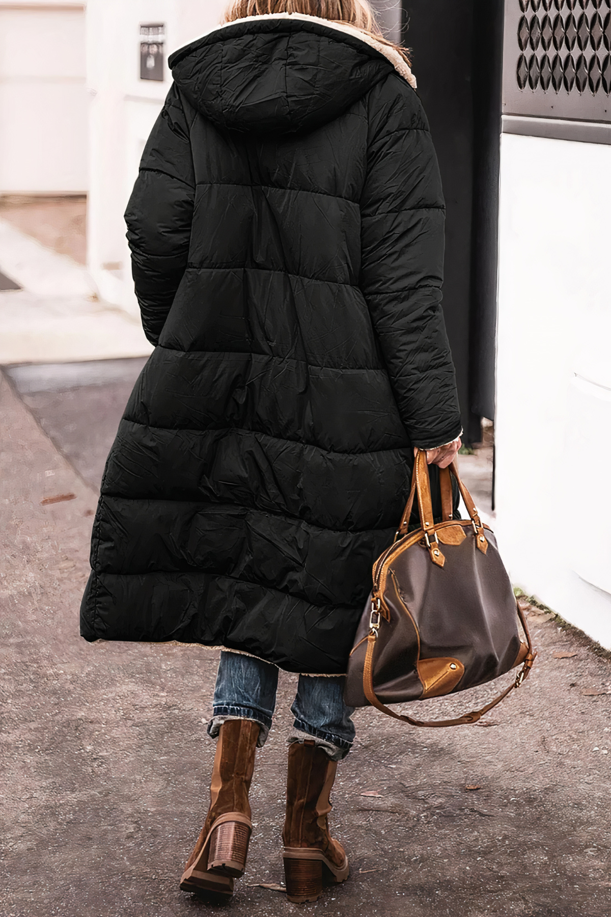 Person wearing a long black puffer coat with a hood, holding a brown leather handbag on a street.