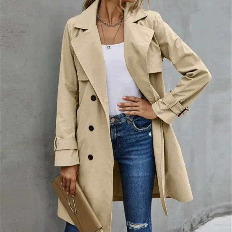 Person wearing a beige trench coat over a white top and blue jeans against a gray background