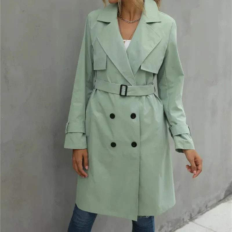 Person wearing a light green trench coat with black buttons against a gray wall.