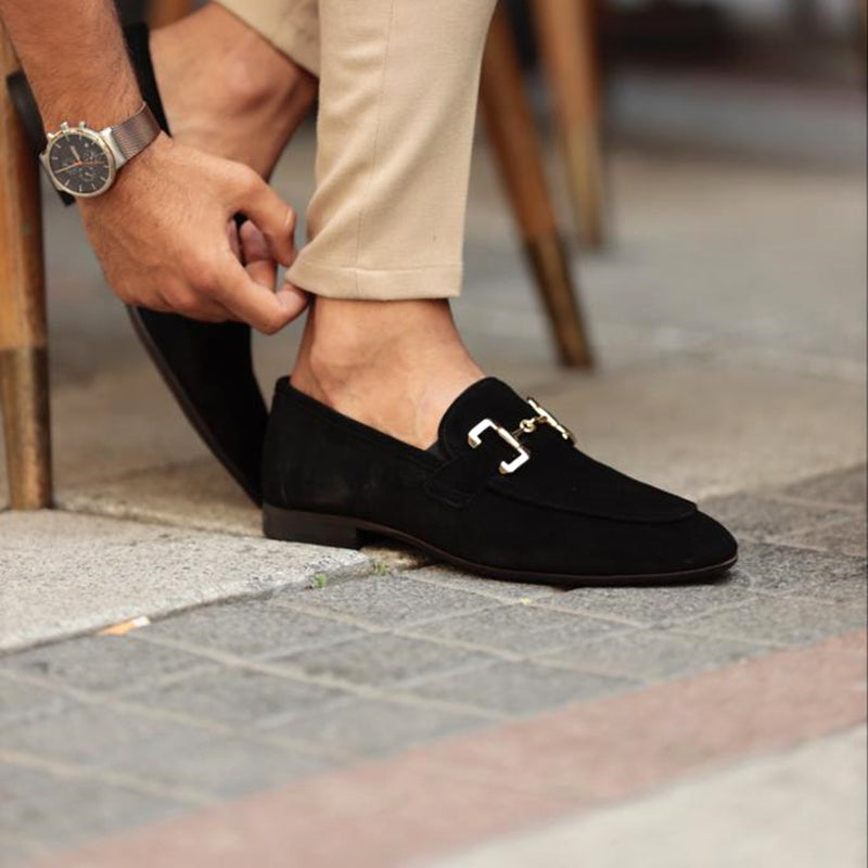 Person wearing black loafers with a gold buckle on a tiled floor.
