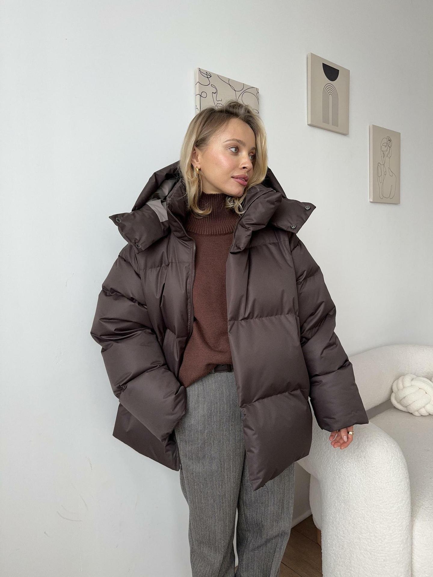Person wearing a brown puffer jacket in a room with a white wall and a white couch.