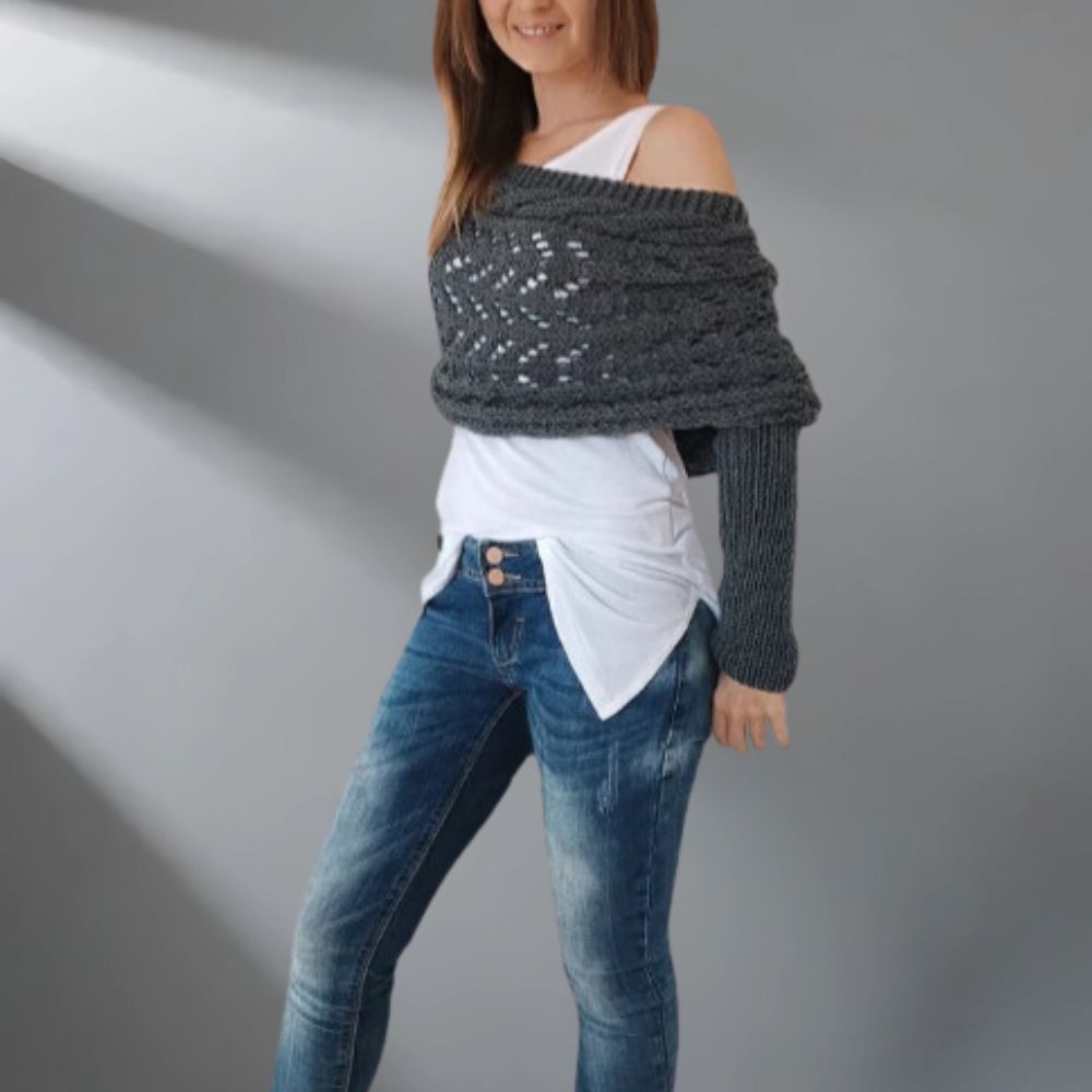 Woman wearing a dark gray knitted shawl over a white top and blue jeans on a plain background