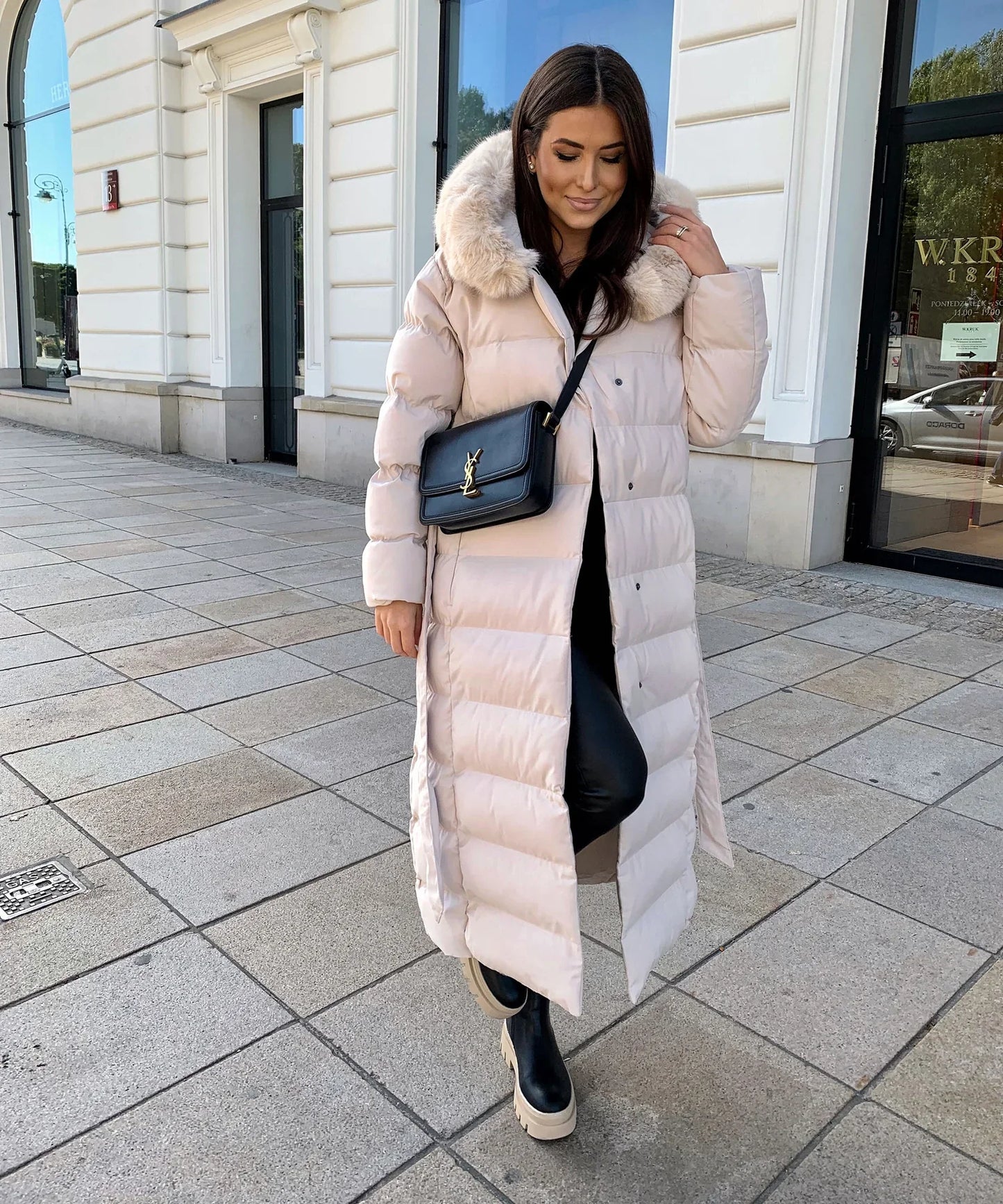 Woman wearing a long beige puffer coat with a fur hood on a city street.