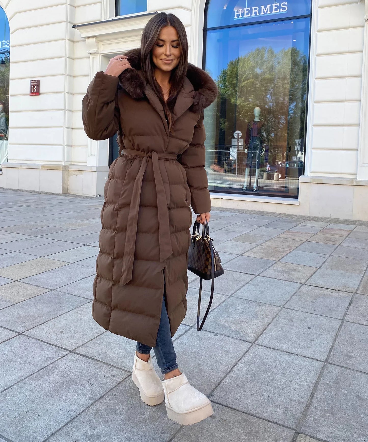 Woman wearing a brown puffer coat in an urban setting with a visible Hermes store.