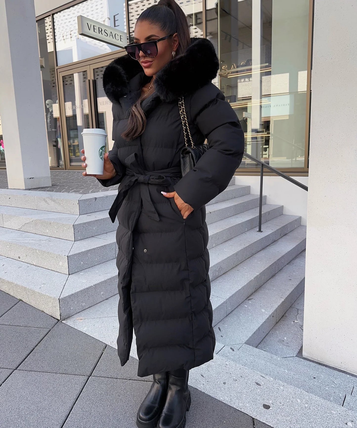 Woman in a black puffer coat holding a Starbucks cup, standing on steps in front of a Versace store.