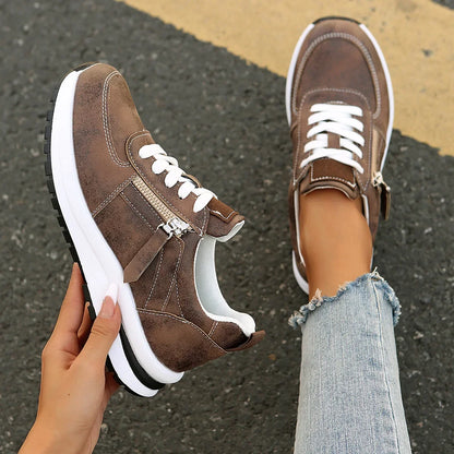 Brown sneakers with white soles held by a hand on a road surface.