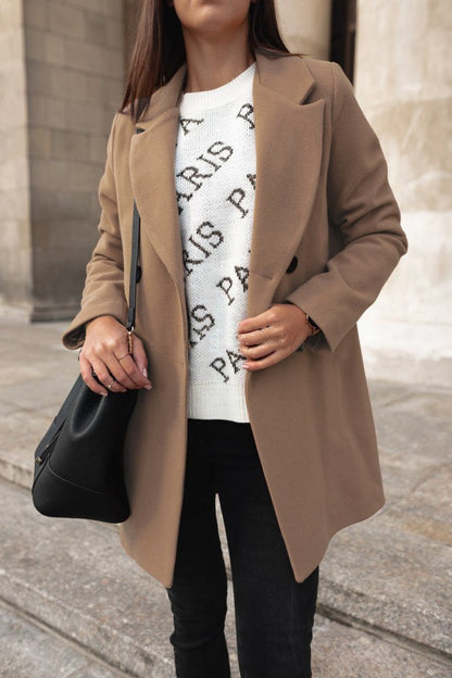Person wearing a beige coat over a white sweater with black text, holding a black handbag.